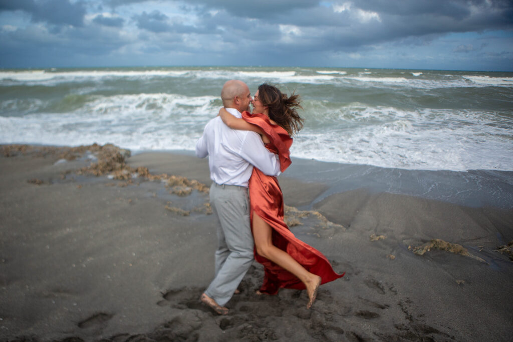 Couple dancing and embracing on rocky shoreline during romantic engagement session at Blowing Rocks Preserve in Florida.