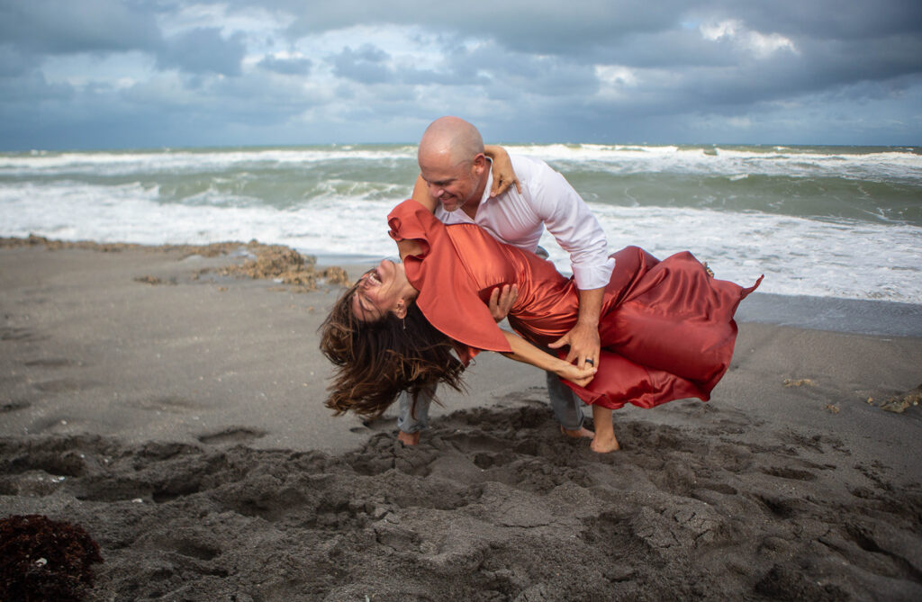 Man dipping laughing woman while dancing during playful engagement session on the beach at Blowing Rocks Preserve, Florida.