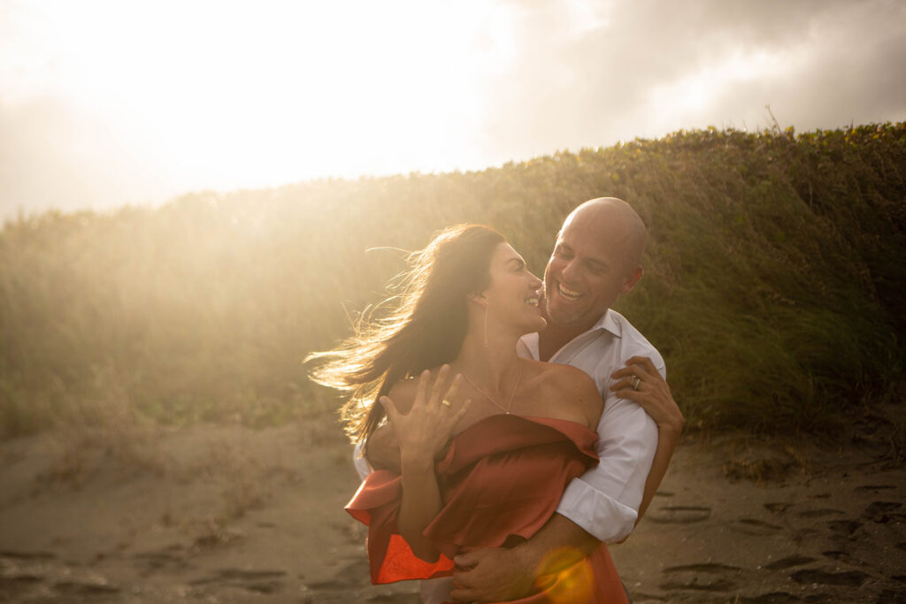 Couple hugging on the beach with woman’s engagement ring clearly visible during intimate engagement session at Blowing Rocks Preserve, Florida.