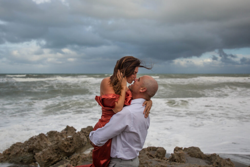 Man lifting woman while she kisses his nose during playful and romantic engagement session at Blowing Rocks Preserve, Florida.