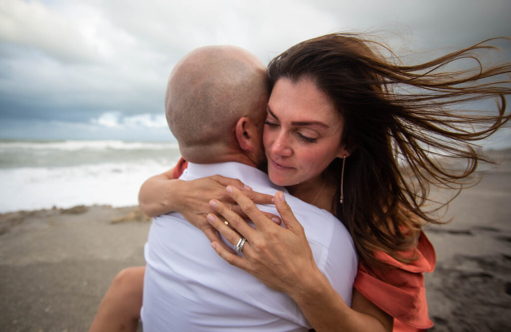 Emotional engagement photo of woman sitting on partner’s lap with legs wrapped around him, crying during intimate beach moment at Blowing Rocks Preserve, Florida.