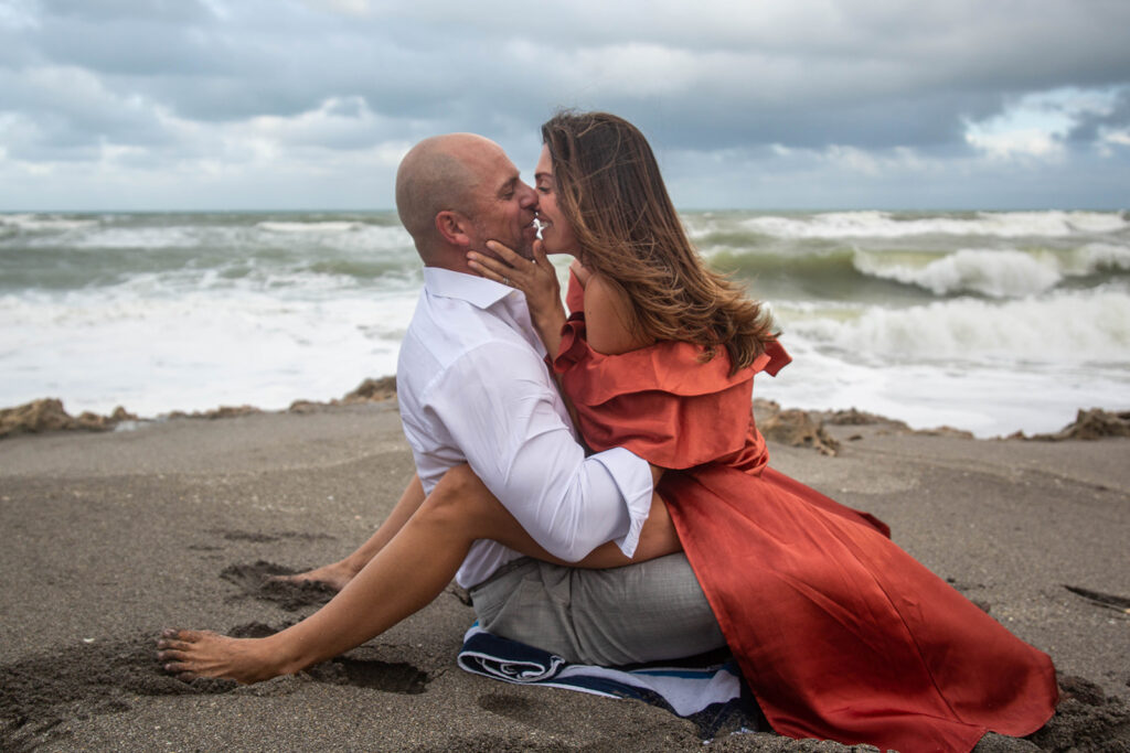 Couple sitting close on the beach with faces nose to nose and smiling, during romantic engagement session at Blowing Rocks Preserve, Florida.