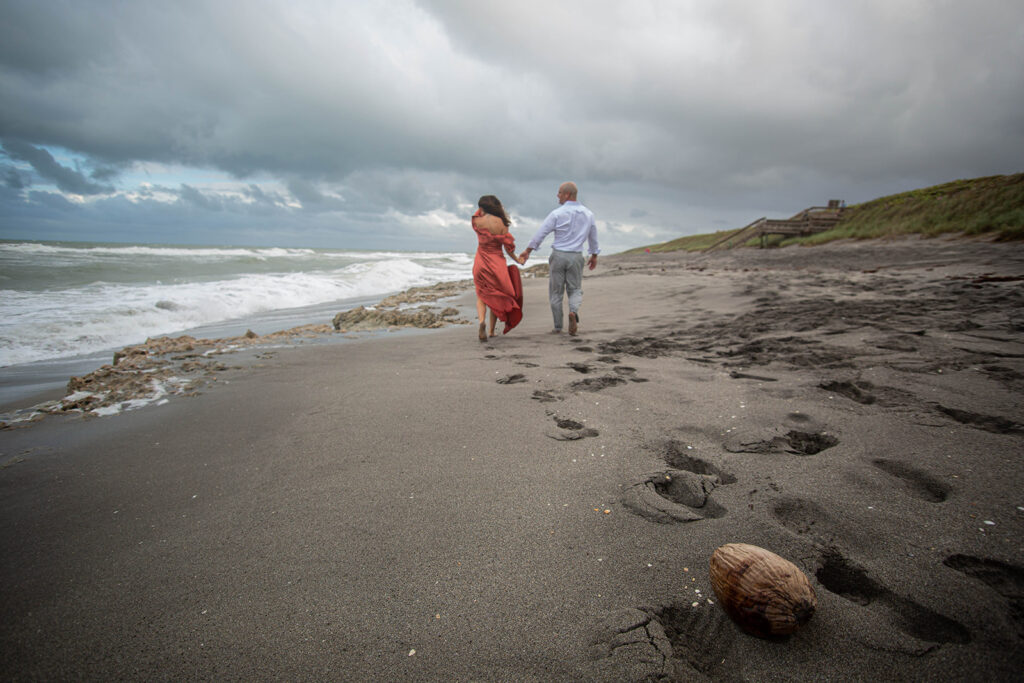 Couple walking hand in hand on the beach with their backs to the camera, footprints in sand in focus at Blowing Rocks Preserve, Florida.