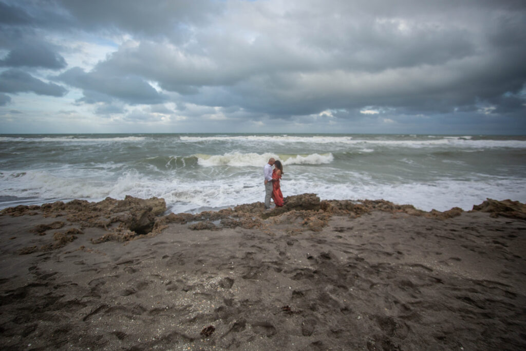 Wide-angle photo of couple embracing on rocky shoreline with ocean backdrop during cinematic engagement session at Blowing Rocks Preserve, Florida.