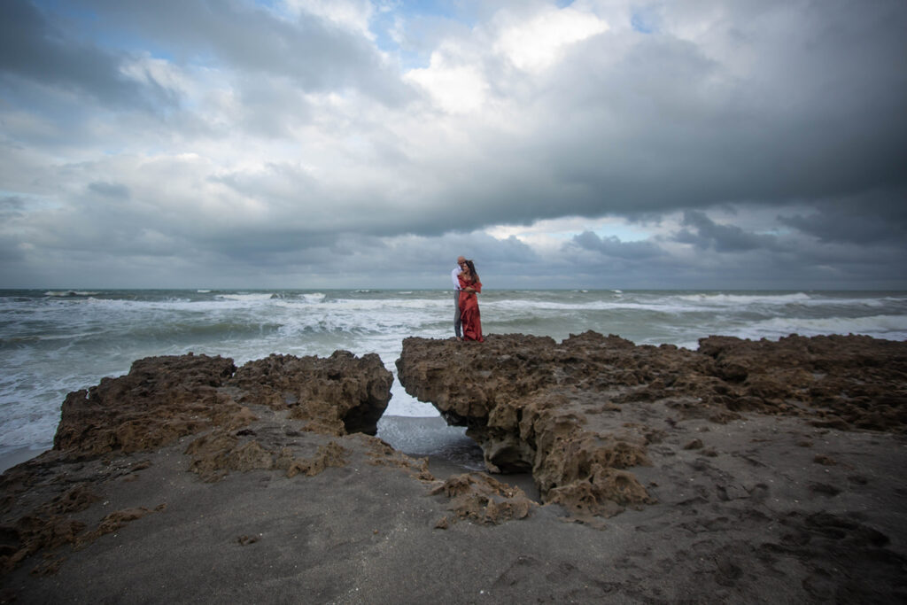 Wide-angle photo of couple on rocky shoreline at Blowing Rocks Preserve, man kissing woman’s shoulder as she looks back during engagement session in Florida.