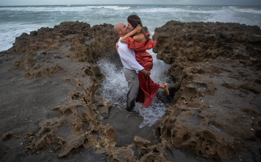 Couple standing on rocky shore with waves rushing in, holding each other close and touching noses while smiling during engagement session at Blowing Rocks Preserve, Florida.