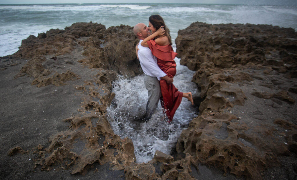 Man lifting fiancée on rocky shoreline as ocean wave crashes behind them during cinematic engagement session at Blowing Rocks Preserve, Jupiter Island, Florida.