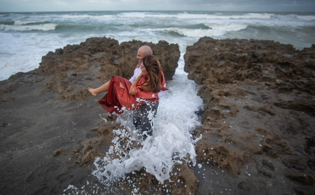 Man helping woman out of a rock hole with waves crashing nearby during engagement session at Blowing Rocks Preserve, Florida.