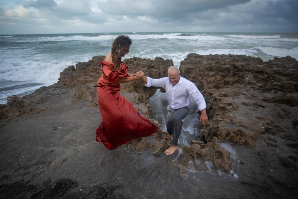 Woman helping man up by holding his hand on rocky shoreline during engagement session at Blowing Rocks Preserve, Florida.