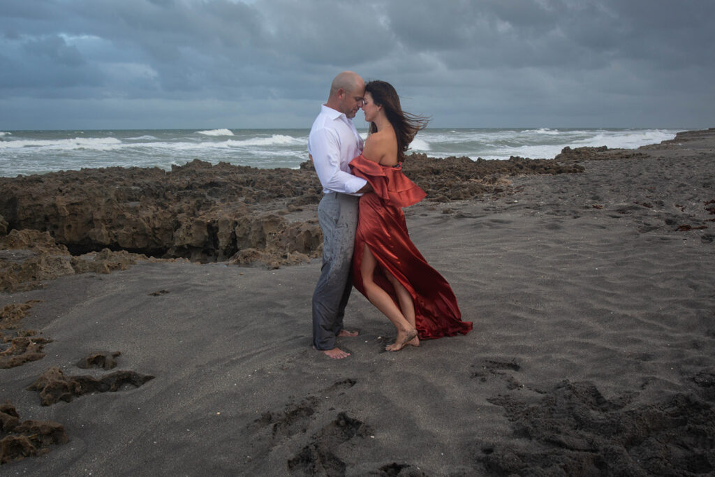 Couple with foreheads touching on rocky shoreline with ocean water behind them during engagement session at Blowing Rocks Preserve, Florida.