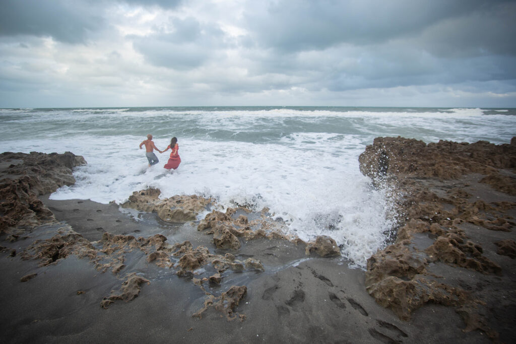 Couple running hand in hand through ocean waves, smiling and laughing during engagement session at Blowing Rocks Preserve, Florida.