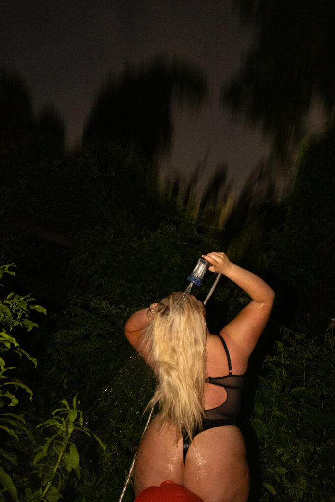 Blonde woman in a black bodysuit lingerie leaning on a red inflatable fire hydrant while spraying water from the hose onto her hair, creating a playful and sexy boudoir image.