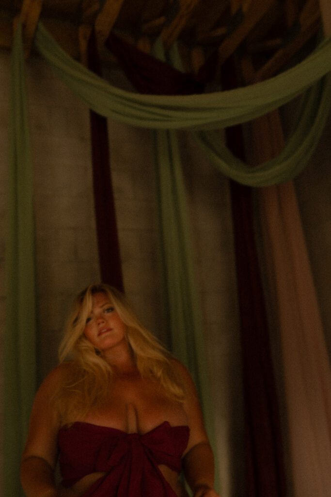 Blonde woman wearing flowing drapes as fabric, staring directly into the camera with a longing, sensual expression, creating a powerful boudoir portrait.