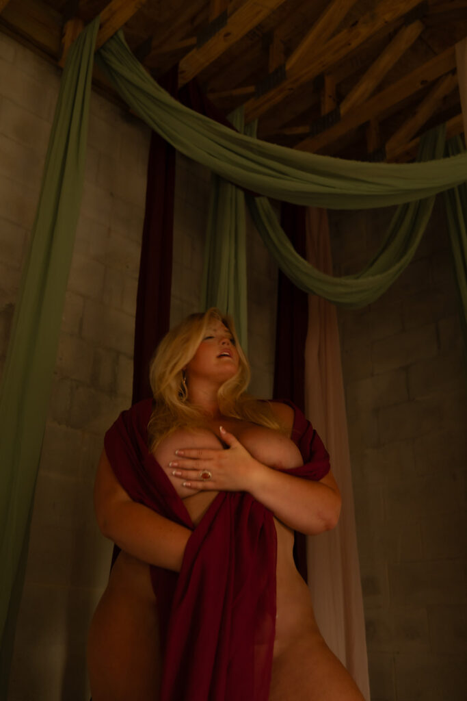 Blonde woman draped in flowing fabric, her body softly revealed as she closes her eyes and turns her head aside, creating a dreamy and sensual boudoir portrait.