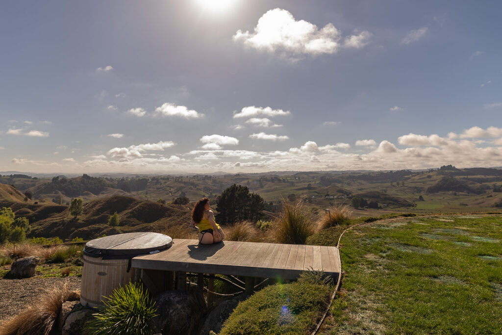 Cecelia poses confidently on a wooden deck in Te Kuiti, New Zealand, playfully holding her hip while showcasing self-assured, bold energy against a mountain backdrop.