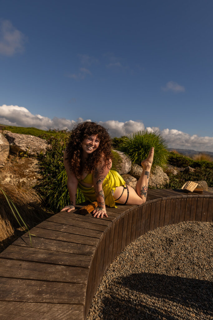 Cecelia smiles at the camera while holding an upward dog pose on a wooden deck in Te Kuiti, New Zealand, with mountain views in the background, blending strength and serenity.