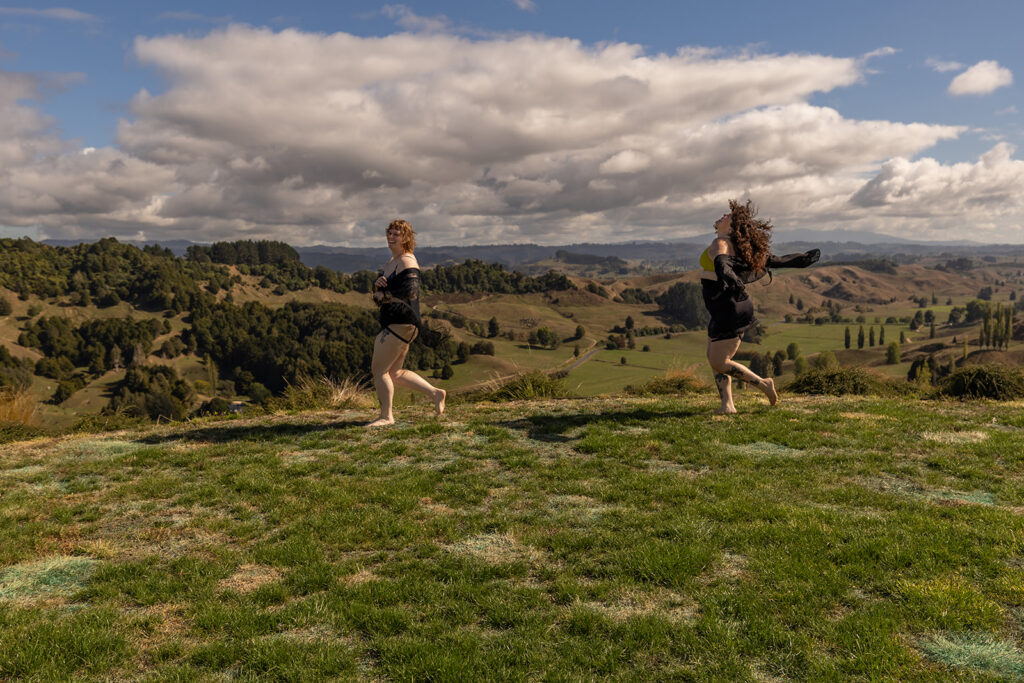 Cecelia and her best friend run and laugh together on a wooden deck surrounded by nature in Te Kuiti, New Zealand, capturing a carefree, joyful moment during their boudoir adventure.