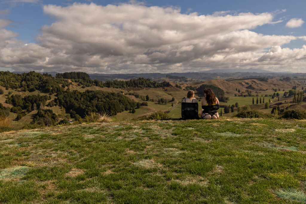 Cecelia and her best friend sit together on a wooden deck overlooking the mountains in Te Kuiti, New Zealand, peacefully admiring the scenic view during their destination boudoir trip.
