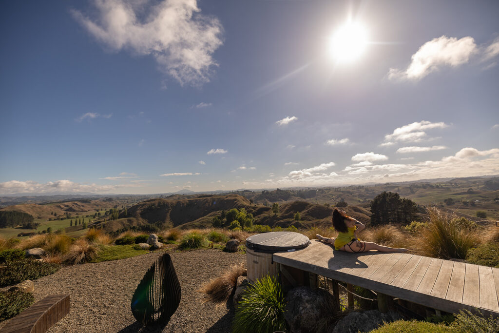 Cecelia sits in a straddle pose on a wooden deck overlooking the mountains in Te Kuiti, New Zealand, capturing a serene and empowering travel moment.