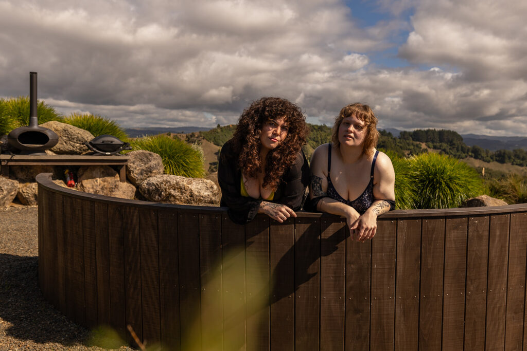 Cece and her best friend sit side by side on a wooden bench in Te Kuiti, New Zealand, looking directly into the camera with bold, confident expressions during their boudoir shoot.