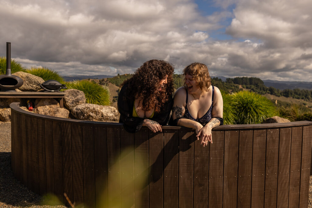 Cecelia and her best friend sit on a wooden bench in Te Kuiti, New Zealand, looking at each other and laughing, capturing a candid moment of joy and friendship during their boudoir adventure.