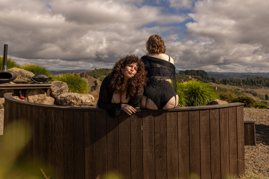 Cecelia makes a playful kissing face toward the camera while her friend Nicole sits on top of a wooden bench in Te Kuiti, New Zealand, capturing a fun and confident moment during their boudoir adventure.