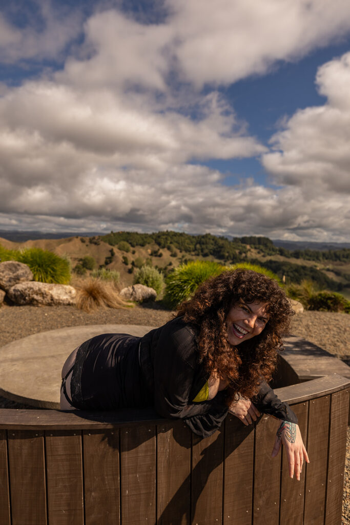 Cecelia leans over a wooden bench in Te Kuiti, New Zealand, laughing joyfully during her destination boudoir shoot, capturing a candid and carefree moment surrounded by nature.