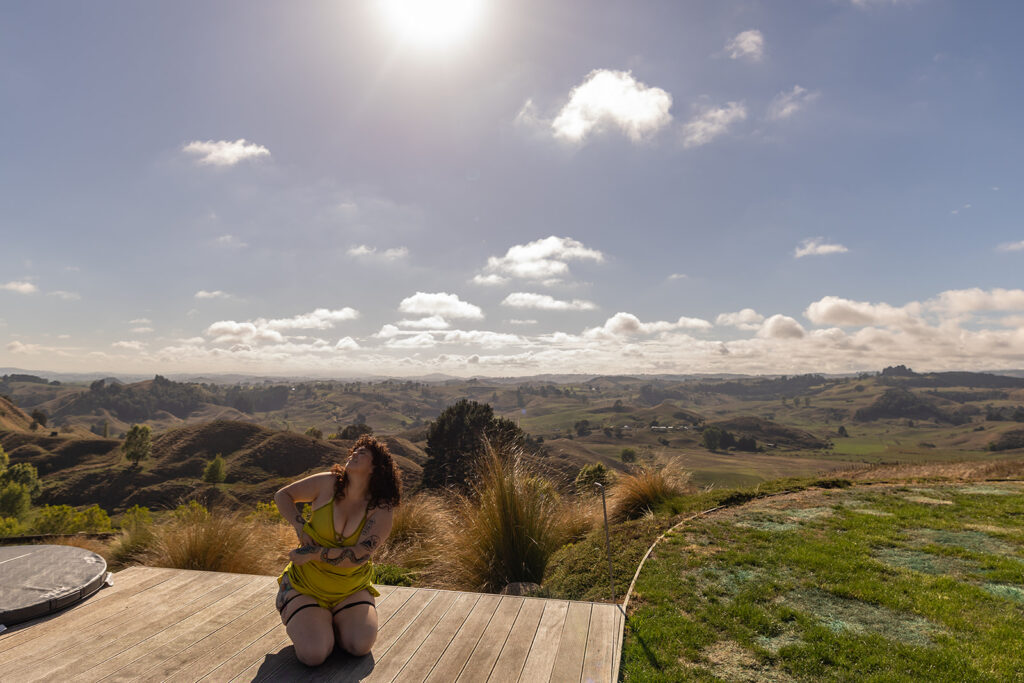 Cecelia gracefully slips one arm out of her dress while standing on a wooden deck in Te Kuiti, New Zealand, capturing a moment of feminine confidence and self-expression against a scenic mountain backdrop.