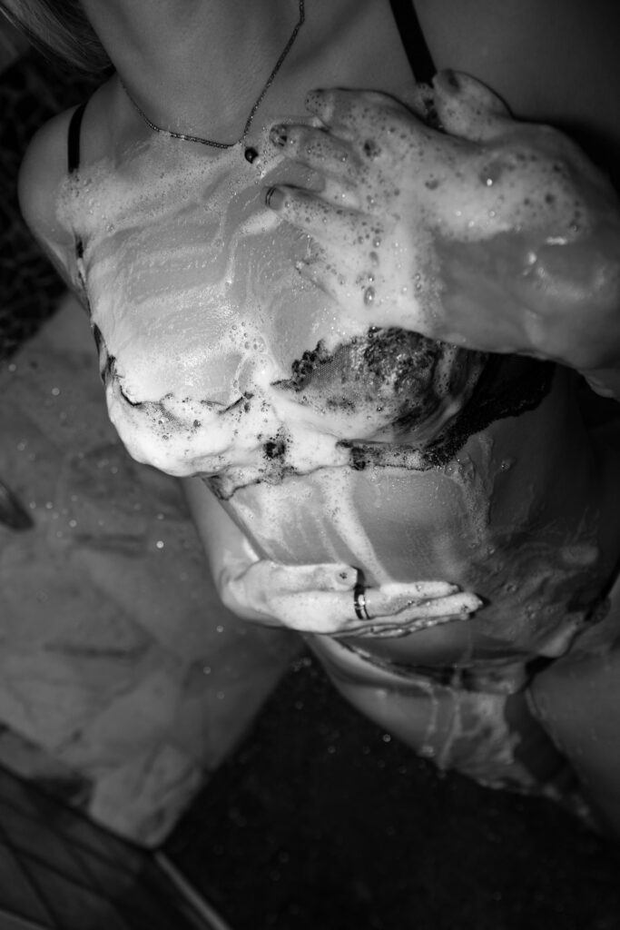 Black-and-white boudoir portrait of a woman in a shower, surrounded by water and soap suds, captured in a cinematic fine art style with soft contrast and texture.